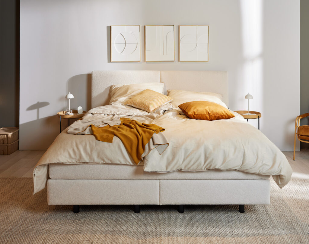 Modernes Schlafzimmer mit neutraler Farbpalette: großes Bett mit beigefarbener Bettwäsche, Senf-gelber Tagesdecke und passenden Kissen, flankiert von zwei weißen Nachttischen mit Tischlampen. An der Wand drei abstrakte Kunstwerke. Senffarbener Sessel und gewebter Teppich ergänzen die minimalistische Einrichtung. Natürliches Tageslicht fällt durch ein Fenster, erzeugt eine ruhige Atmosphäre