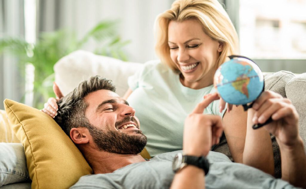 Zwei Personen befinden sich in einem gemütlichen Wohnzimmer. Eine Person liegt entspannt auf einem Sofa mit einem gelben Kissen, während die andere neben dem Sofa sitzt und einen kleinen Globus hält. Die sitzende Person zeigt auf eine Stelle auf dem Globus, möglicherweise diskutieren sie eine Reise. Im Hintergrund sind eine grüne Pflanze und ein Fenster mit Vorhängen zu sehen.