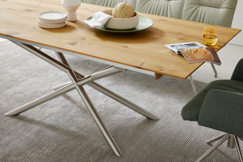Modern gedeckter Esstisch mit Holzplatte und schwarzen Metallbeinen. Auf dem Tisch stehen gestapelte weiße Teller, eine helle Keramikvase, ein grüner Keramikbehälter mit einer Melone, ein zusammengelegtes weißes Tuch, ein Glas mit gelbem Getränk und ein Magazin mit der Aufschrift „FASHION“. Der Tisch steht auf einem strukturierten grauen Teppich. Zwei Stühle – einer in Grün, einer in Beige – flankieren den Tisch. Die Szene wirkt stilvoll und zugleich gemütlich, ideal für ein entspanntes Essen im zeitgemäßen Wohnbereich.