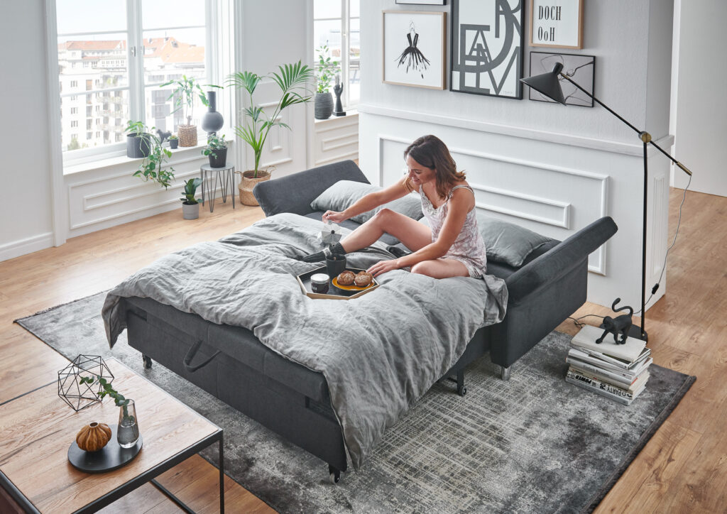 Modernes Schlafzimmer mit grauem Schlafsofa, Frühstückstablett und Pflanzen – stilvolle Einrichtung für entspannte Morgenstunden mit gemütlichem Flair und natürlichem Licht.