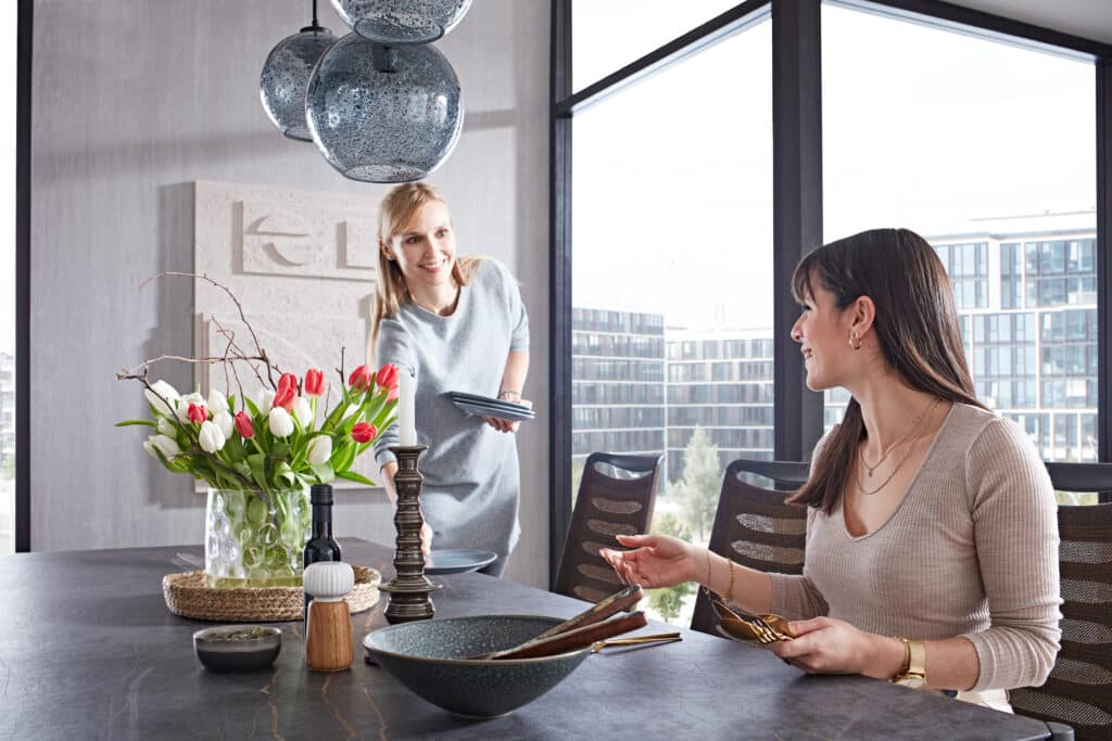 Modernes Esszimmer mit zwei Personen, die den Tisch decken. Auf dem Holztisch stehen ein Tulpenstrauß in Rot und Weiß, ein Kerzenhalter mit weißer Kerze, Salz- und Pfefferstreuer, eine Schüssel mit Essstäbchen und ein kleines Schälchen. Über dem Tisch hängen drei kugelförmige Pendelleuchten aus strukturiertem Glas. Im Hintergrund große Fenster mit Blick auf moderne Gebäude.