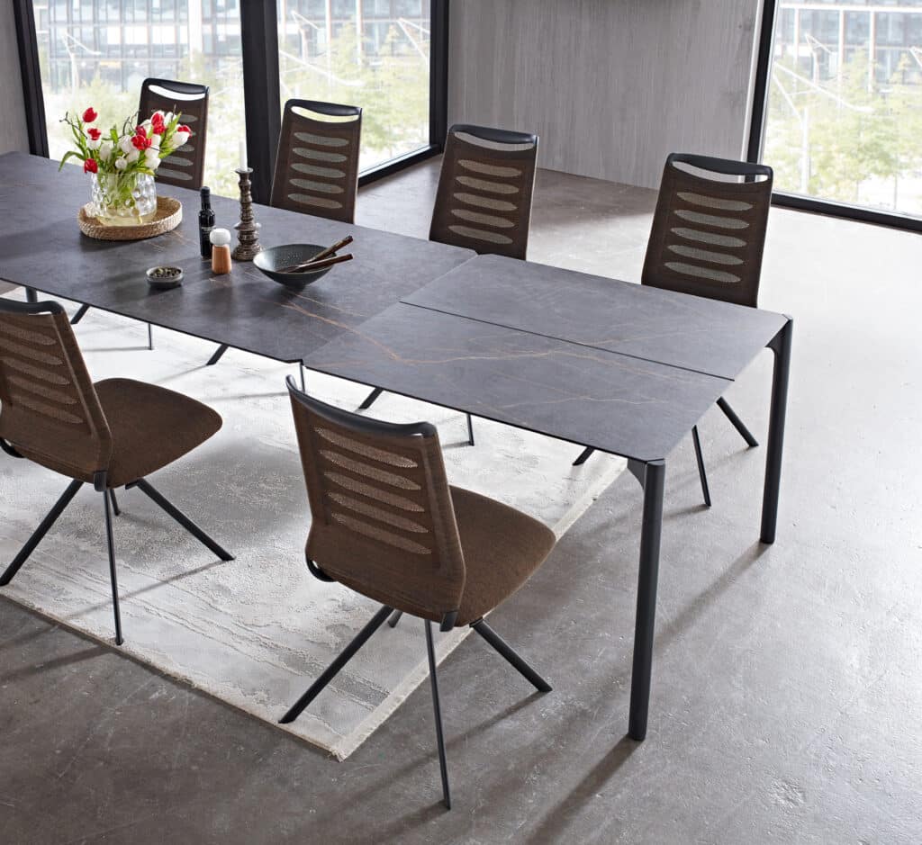 Modernes Esszimmer mit rechteckigem Tisch in dunkler Steinoptik und sechs braunen Stühlen mit gepolstertem Sitz und Netzrücken. Auf dem Tisch steht ein Glasvase mit roten und weißen Blumen auf einem geflochtenen Tischset, daneben ein schwarzer Teller, eine kleine Schale, Salz- und Pfefferstreuer. Der Raum hat große Fenster mit Blick auf ein Gebäude und Bäume, der Boden ist aus Beton mit einem hellen Teppich unter dem Tisch.