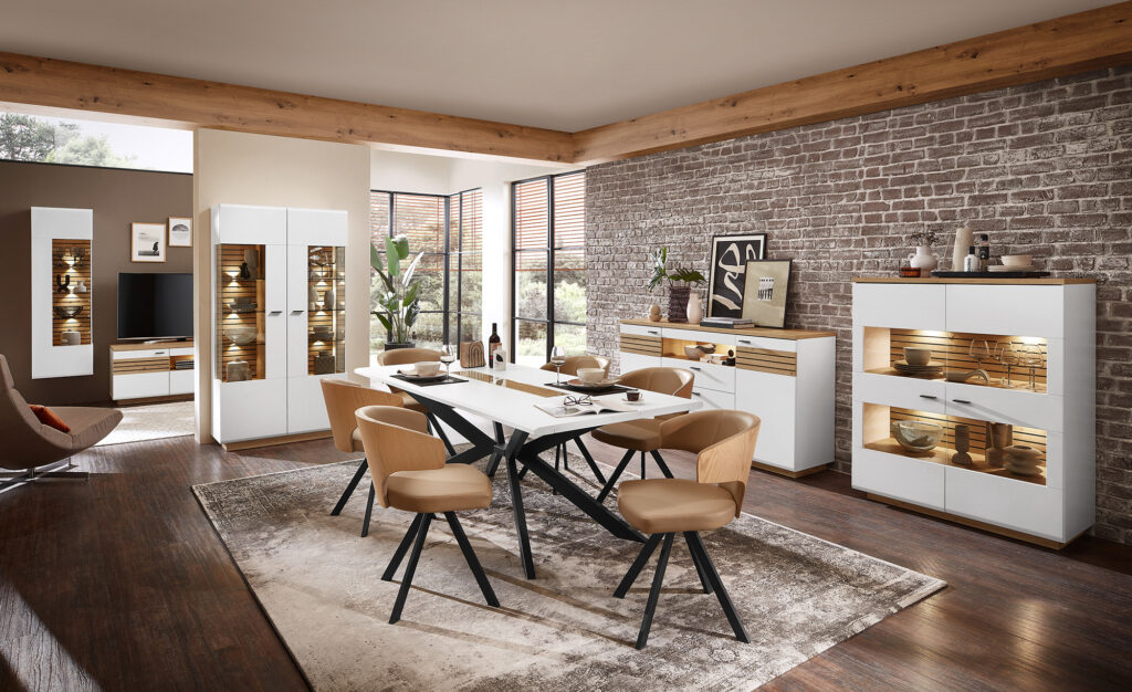 Stilvoll eingerichtetes Wohnzimmer mit heller, moderner Einrichtung. Im Zentrum steht ein großer weißer Esstisch mit schwarzem Sockel, umgeben von sechs beigen Stühlen mit Holzdetails. Umgeben ist der Raum von weißen Möbelstücken mit Glas- und Holzakzenten, darunter ein Schrank, ein Sideboard und ein TV-Regal mit Fernseher. Die Wände sind teils weiß gestrichen, teils mit Ziegeloptik gestaltet, und es hängen gerahmte Bilder. Große Fenster lassen viel Tageslicht herein, ergänzt durch sichtbare Holzbalken an der Decke. Pflanzen, Vasen und Bücher sorgen für wohnliche Atmosphäre.