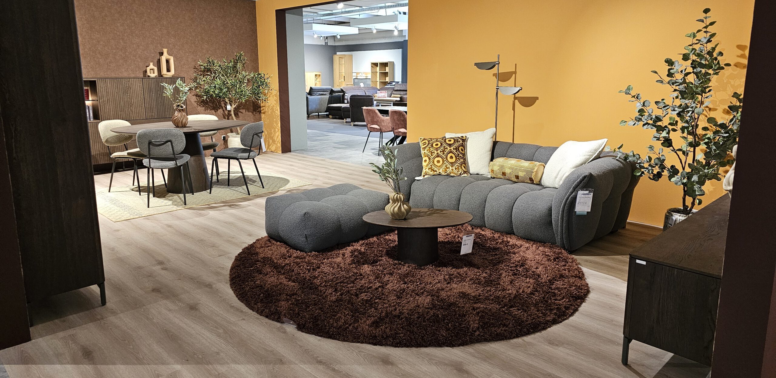 Moderne Wohn- und Esszimmerausstellung im Möbelhaus mit grauem Ecksofa, rundem Teppich, stilvoller Beleuchtung, rundem Esstisch und warmen Wandfarben – gemütliche und trendbewusste Einrichtung im XOOON-Stil.
