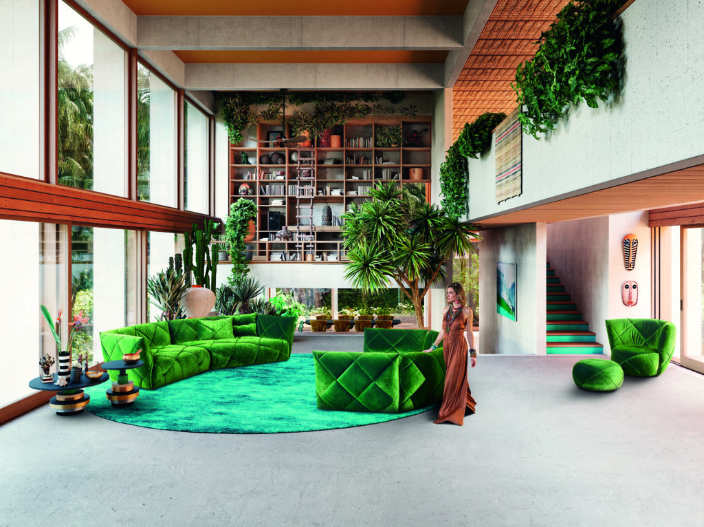 Wohnzimmer mit grüner Bretz-Sitzgruppe auf blauem Teppich, umgeben von Pflanzen, Kunstobjekten und einer Bücherwand – farbenfrohes, modernes Interior mit lebendiger Atmosphäre.