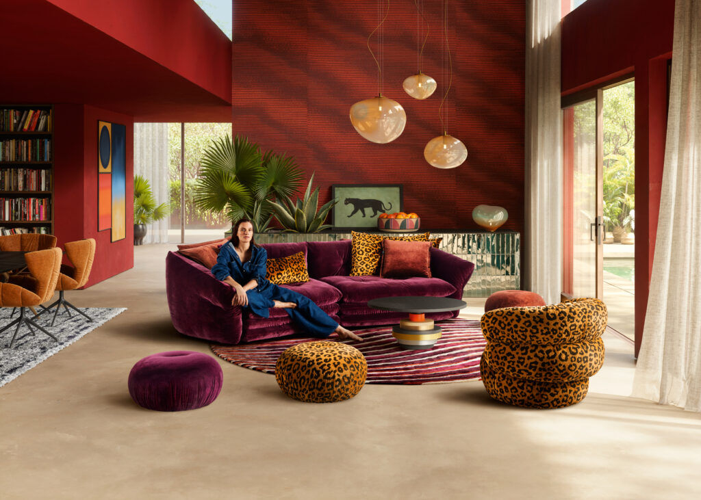 Modernes Wohnzimmer mit hoher Decke und großen Fenstern, roter Wandgestaltung, lilafarbener Samtcouch mit gemusterten Kissen, rundem Couchtisch auf gestreiftem Teppich, Leopardenhockern und Pflanzen – stilvoll und farbenfroh mit Blick ins Grüne.