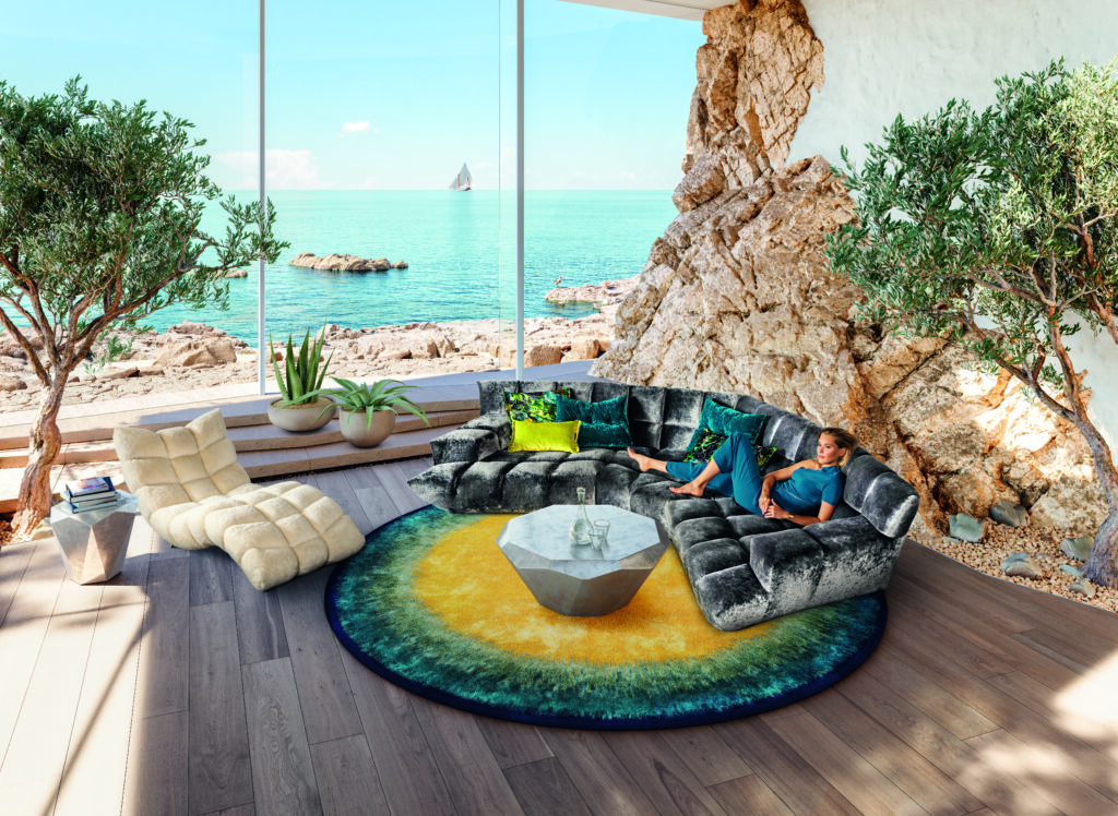 Modernes Wohnzimmer mit großem Panoramafenster und Meerblick, grauer Eckcouch mit bunten Kissen, rundem Farbverlauf-Teppich, geometrischem Couchtisch, Lounge-Sessel und mehreren Zimmerpflanzen – stilvolle Einrichtung mit natürlichem Übergang zur Felslandschaft draußen.