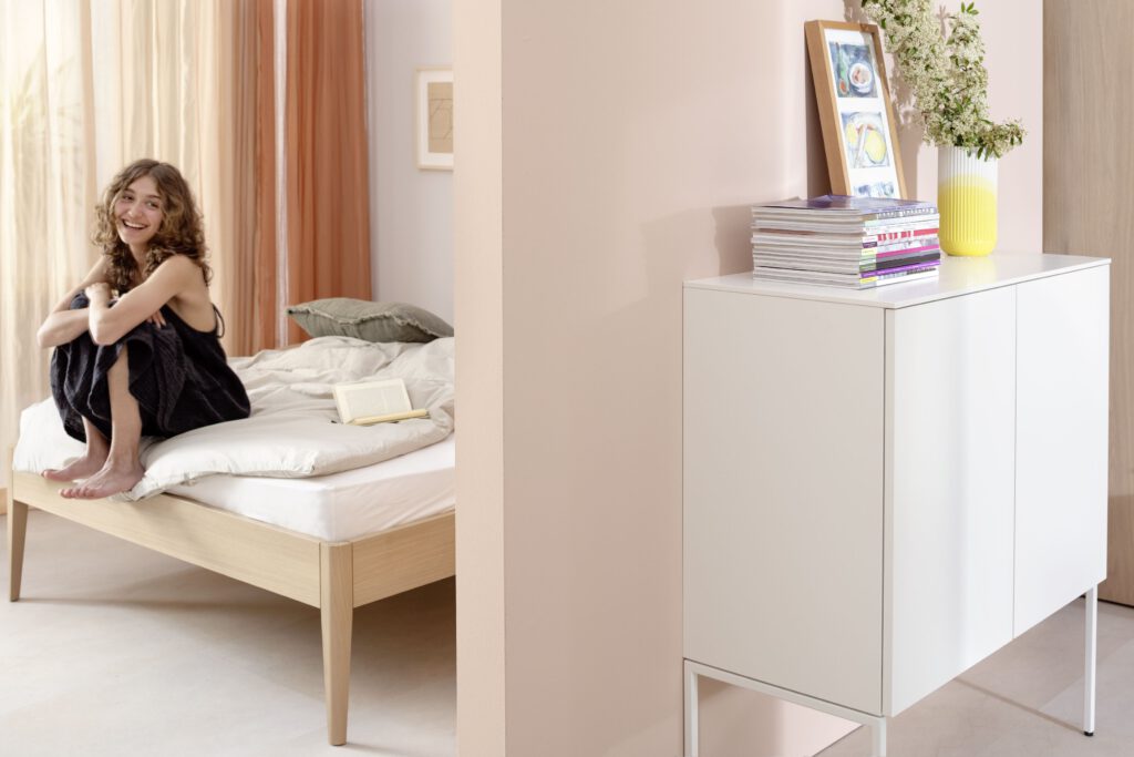 Modernes Schlafzimmer mit Holzbett, weißer Bettwäsche und ruhiger Farbgestaltung. Rechts ein weißes Sideboard mit Vase, Büchern und Wandbild