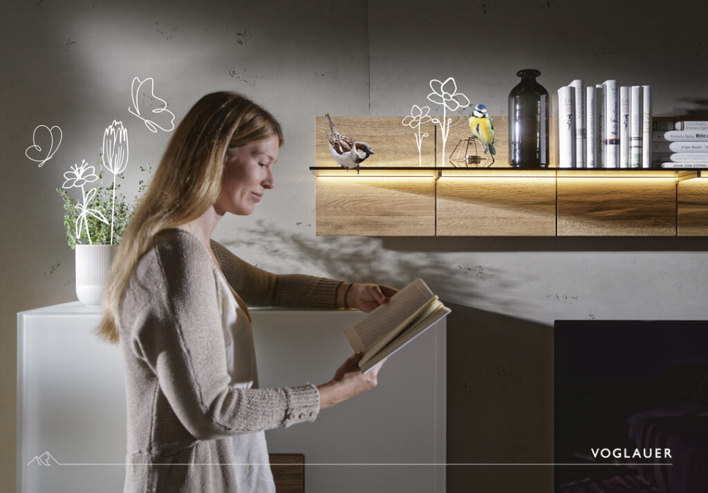 Frau liest ein Buch neben einem weißen Sideboard mit Pflanze; darüber ein beleuchtetes Holzregal mit Deko und Büchern; Wand mit floralen Illustrationen; Voglauer-Möbel.