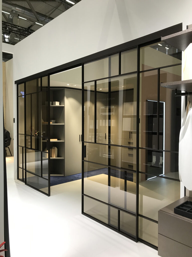 Modernes Interieur mit schwarzen Gleittürprofilen und Glas-Raumteilern, vermutlich aus dem Raumplus Gleittürsystem S720. Die rahmenlosen Glaspaneele schaffen eine elegante, transparente Abtrennung in einem minimalistischen Raum mit weißen Böden und sichtbaren Industrieelementen an der Decke. Im abgetrennten Bereich befinden sich Regale mit dekorativen Objekten – ideal für Büro, Showroom oder modernes Wohnen.
