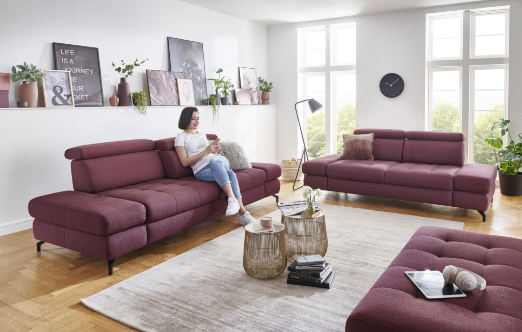Modernes Wohnzimmer mit zwei weinroten Sofas und passendem Hocker, goldfarbenen Beistelltischen, Pflanzen, Wanddeko und großem Fenster. Stilvoll und gemütlich eingerichtet