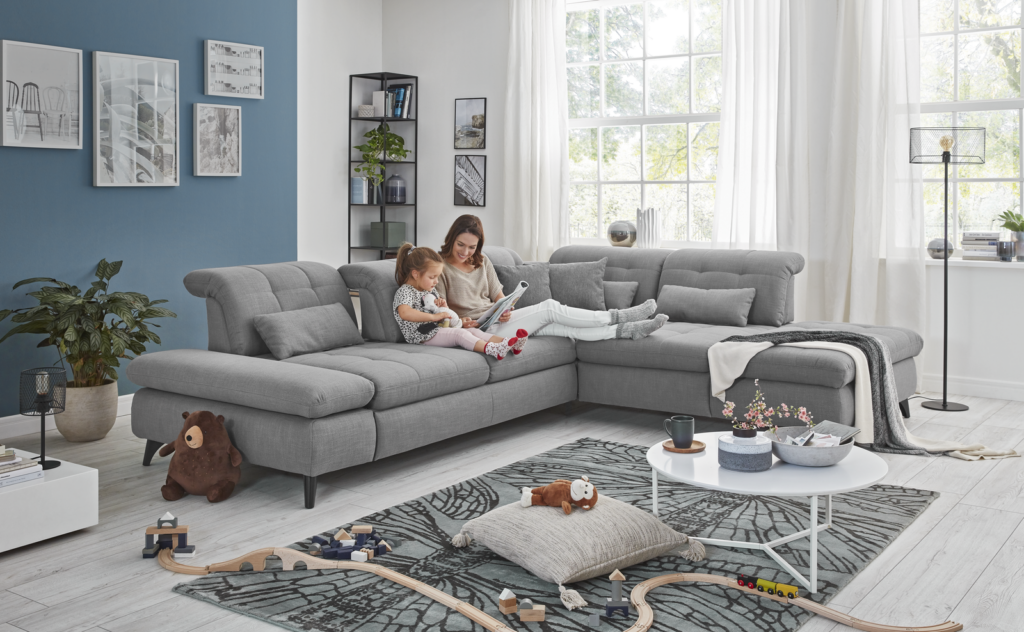 Modernes Wohnzimmer mit großer grauer Eckcouch, auf der zwei Personen gemeinsam ein Buch lesen. Der Raum hat eine blaue Akzentwand mit gerahmten Bildern, ein schwarzes Regal mit Büchern und Pflanzen, ein großes Fenster mit weißen Vorhängen sowie einen weißen Couchtisch mit Deko. Auf dem Boden liegen eine Spielzeugeisenbahn und Kuscheltiere, eine grüne Zimmerpflanze steht neben dem Sofa