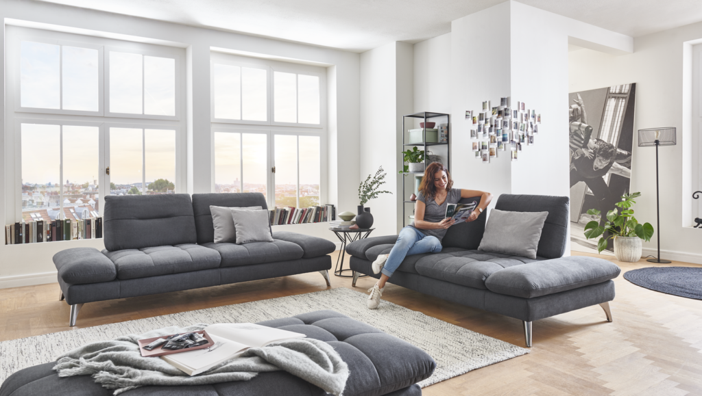 Modernes Wohnzimmer mit zwei dunkelgrauen Sofas, hellem Teppich und kleinem Couchtisch mit Pflanze. Eine Person sitzt lesend auf dem Sofa, hinter ihr eine Fotocollage an der Wand. Große Fenster, Bücherregal, Stehlampe und Zimmerpflanze sorgen für eine helle und gemütliche Atmosphäre