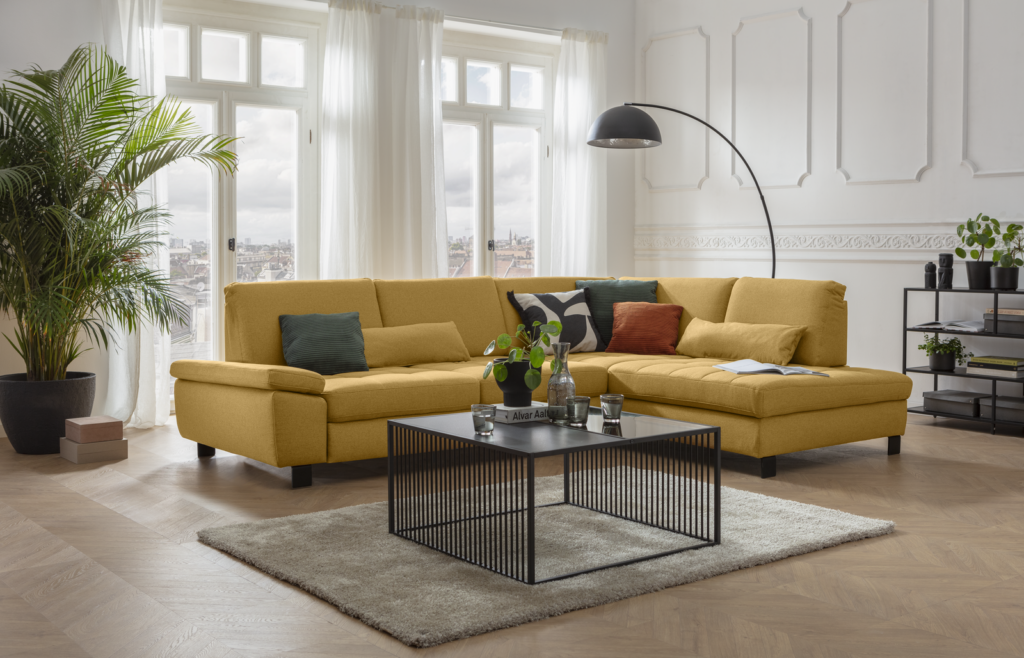 Modernes Wohnzimmer mit großer gelber Eckcouch und bunten Kissen. Schwarzer Couchtisch mit Glasplatte steht auf hellgrauem Teppich, dekoriert mit Pflanze, Buch und Glasgefäßen. Links große Zimmerpflanze vor weißen Fenstern mit transparenten Vorhängen, rechts schwarze Stehlampe mit gebogenem Arm. Im Hintergrund Regal mit Deko und Pflanzen, weiße Wände mit Stuck und heller Fischgrät-Holzboden