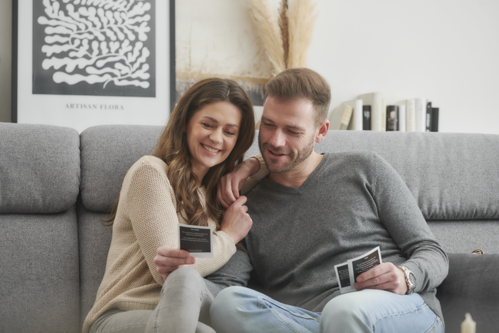 Zwei Personen sitzen auf einem grauen Sofa in einem Wohnzimmer und halten Karten in der Hand, offenbar in ein gemeinsames Gespräch oder Spiel vertieft. Hinter ihnen hängt ein gerahmtes schwarz-weißes Kunstwerk mit der Aufschrift „ARTISAN FLORA“. Auf einem Regal befinden sich dekorative Gegenstände, darunter eine Vase mit Trockenpflanzen und eine Reihe von Büchern. Die Atmosphäre wirkt gemütlich und entspannt.
