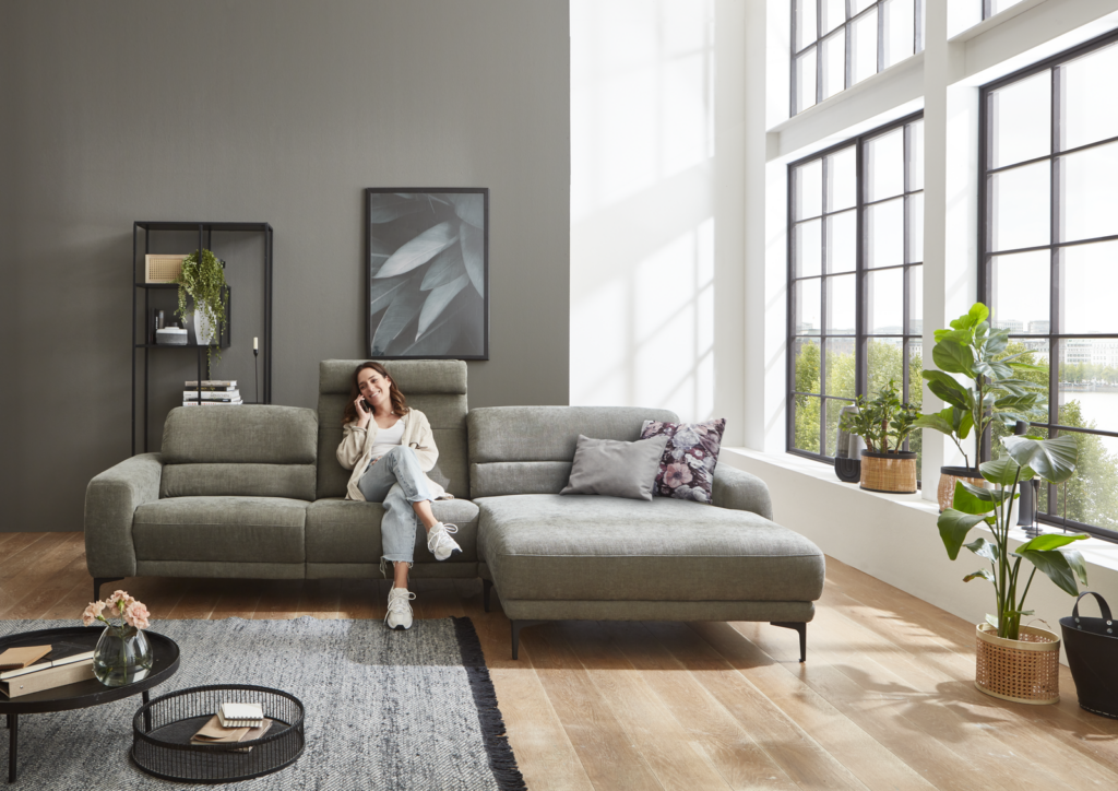 Modernes Wohnzimmer mit einem großen, hellgrauen Ecksofa, das rechts eine Chaiselongue hat. Auf dem Sofa liegen mehrere Kissen, darunter eines mit Blumenmuster. Eine Person sitzt auf dem Sofa und hält ein Smartphone. Der Raum ist durch ein großes Fenster mit schwarzen Rahmen hell erleuchtet. Neben dem Fenster stehen mehrere Zimmerpflanzen. Links im Bild befindet sich ein schwarzes Metallregal mit Dekoration, darunter Bücher und eine Pflanze. Über dem Sofa hängt ein gerahmtes Kunstwerk mit Blattmotiv. Vor dem Sofa steht ein runder Couchtisch mit einer Blumenvase, einem Buch und einem Tablett. Der Boden ist aus hellem Holz, unter dem Tisch liegt ein grauer Teppich.