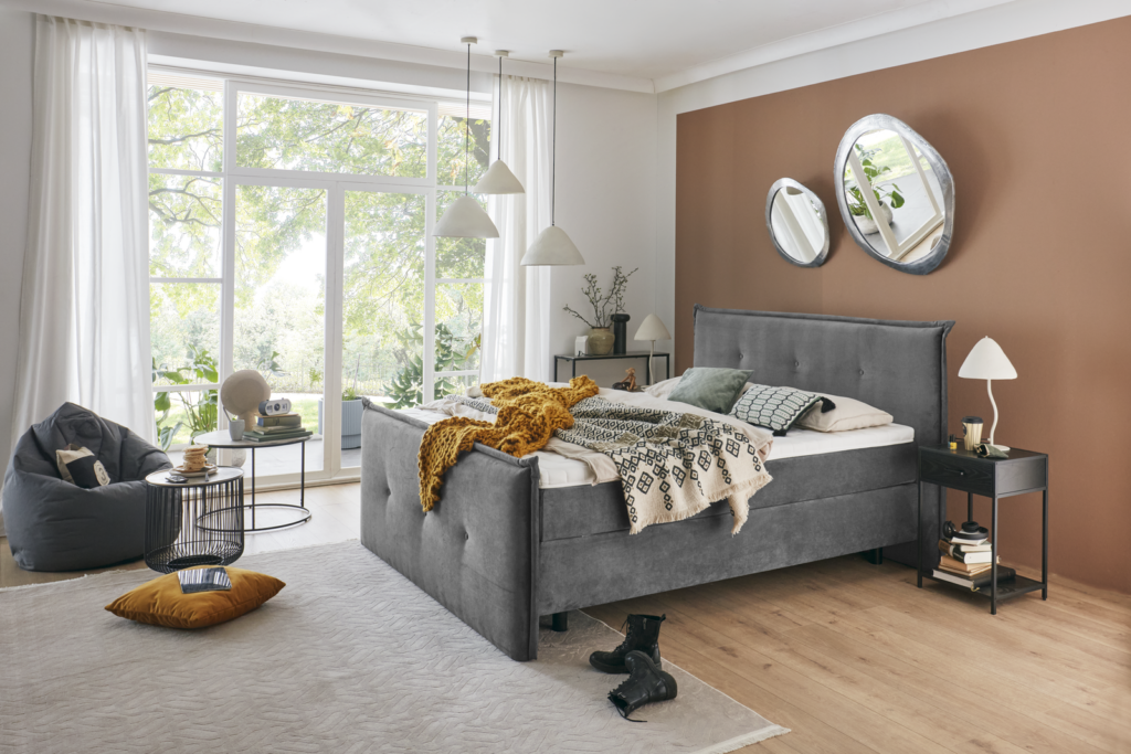 Gemütlich eingerichtetes Schlafzimmer mit grauem Polsterbett, dekorativen Kissen und Decken. Links ein Sitzbereich mit grauem Sitzsack, rundem Tisch, Büchern und Globus. Rechts zwei ovale Wandspiegel über dem Nachttisch. Schwarze Stiefel stehen am Fußende des Betts, große Fenster lassen Tageslicht herein und schaffen eine helle, einladende Atmosphäre