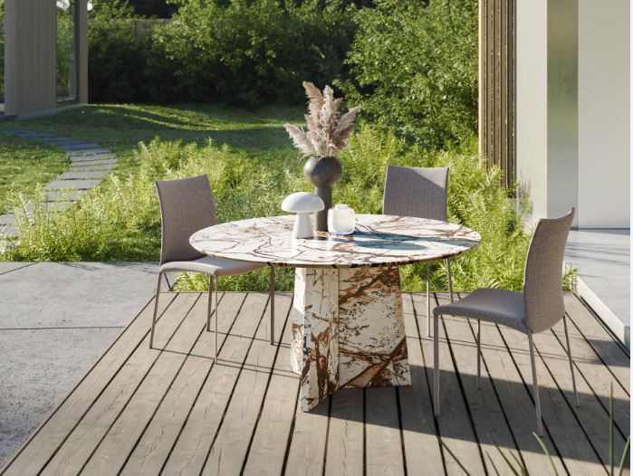Outdoorbereich mit rundem Tisch in Marmoroptik, grauen Stoffstühlen, Pampasgras-Vase und Deko auf Holzdeck vor grüner Gartenkulisse mit Sonnenlicht.