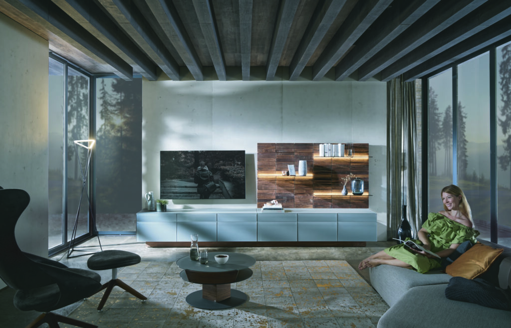 Stilvolles Wohnzimmer mit grauem Sofa, Loungechair und moderner TV-Wohnwand; große Fenster mit Waldblick, Betonwände und warme Beleuchtung schaffen ein elegantes, naturverbundenes Ambiente.