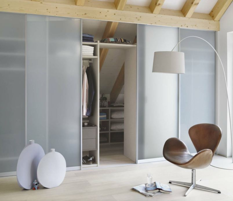 Modernes Wohnambiente mit einem Gleittürenschranksystem, vermutlich Raumplus S720. Der geöffnete Schrank zeigt ordentlich gefaltete Kleidung und hängende Hemden auf Regalböden. Der Raum ist minimalistisch gestaltet mit Holzdeckenbalken, einem schlichten Stuhl, einer Stehlampe und dekorativen Elementen auf dem Boden wie runde Objekte, ein Spielzeug und eine Zeitschrift. Die Szene vermittelt Ordnung, Stil und zeitgemäßes Design.