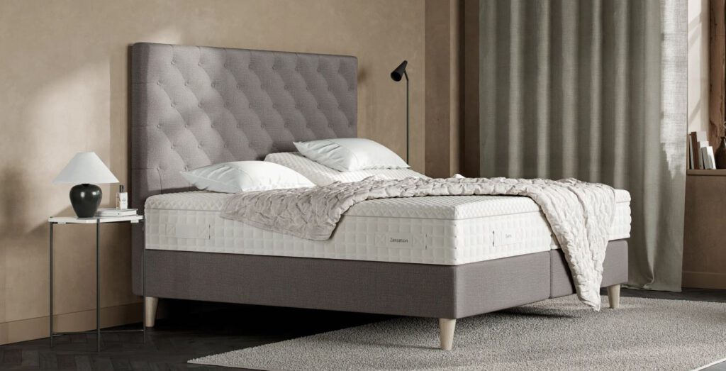 Modernes Schlafzimmer mit Zenstation-Matratze, grauem Kopfteil, weißer Bettwäsche, beiger Decke, stilvollen Möbeln und natürlichem Licht.