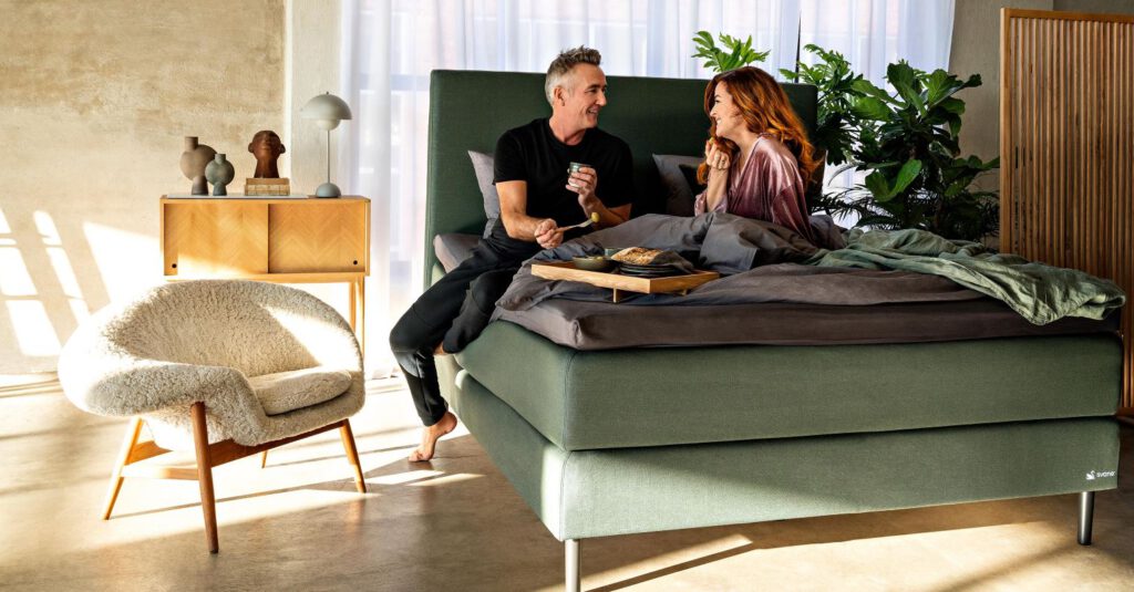 Modernes Schlafzimmer mit grünem Bett, auf dem ein Paar sitzt; Frühstückstablett, stilvolle Einrichtung, Sonnenlicht und Pflanzen schaffen gemütliche Atmosphäre.