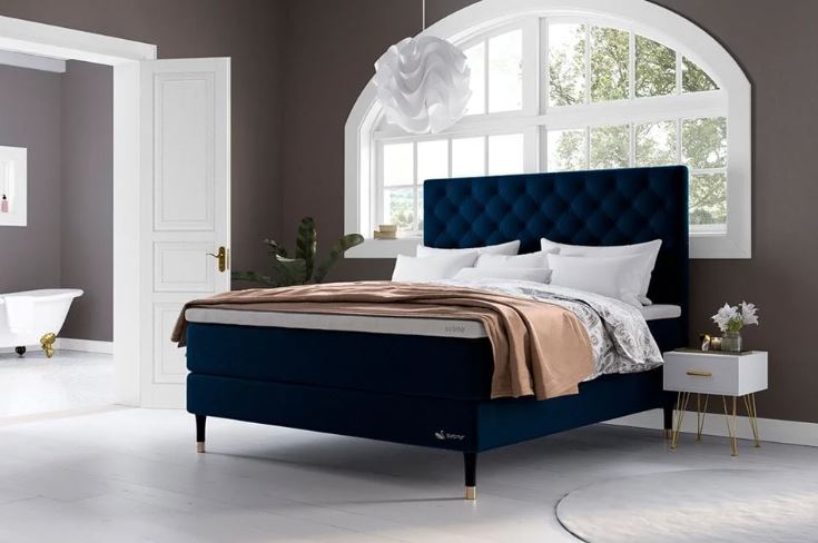 Elegantes Schlafzimmer mit dunkelblauem Polsterbett, weiß-goldenem Nachttisch, großem Fenster, dunklen Wänden und stilvoller, moderner Einrichtung.