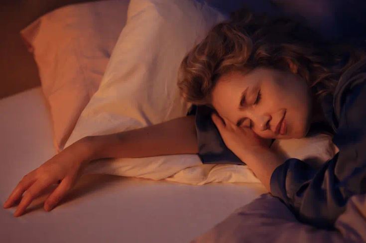 Person liegt entspannt auf einem Bett mit Kissen, in warmem Licht und gemütlicher Atmosphäre, trägt langärmlige Kleidung.