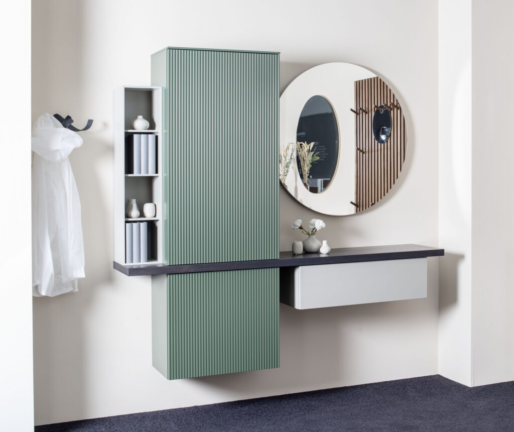 Modernes Badezimmer mit wandmontierter grüner Rillenfront, offenen Regalfächern, dunkler Waschtischplatte und rundem Spiegel – stilvolle Kombination aus Funktion und Design.