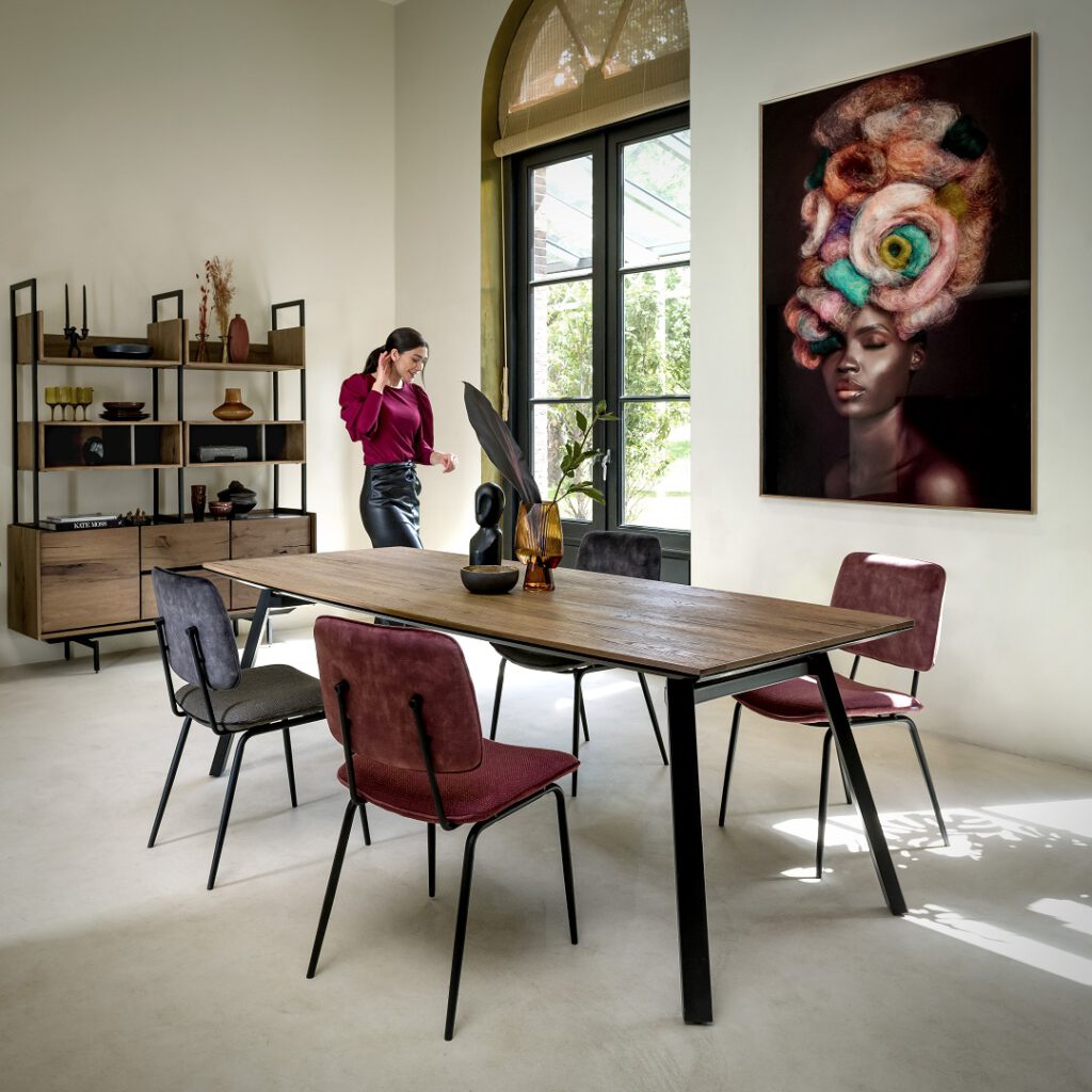 Modernes Esszimmer mit Holz-Tisch und schwarzen Metallbeinen, umgeben von vier Stühlen in Rot und Grau. Auf dem Tisch stehen eine schwarze Skulptur, eine Vase mit Pflanzen und eine Schale. Im Hintergrund ein großes Fenster mit Rundbogen, ein Regal mit Dekoobjekten und ein farbenfrohes abstraktes Gemälde an der Wand.