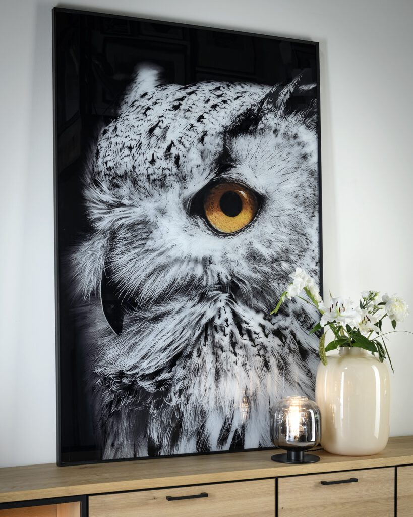 Großformatige Fotografie eines Eulen-Gesichts, mit Fokus auf das leuchtend gelbe Auge und die fein strukturierten Federn. Das Bild ist gerahmt und hängt über einem Holzschrank. Auf dem Schrank stehen eine große beigefarbene Vase mit weißen Blumen sowie ein kleines Glas mit einer brennenden Kerze. Die Komposition wirkt durch das intensive Eulenauge und die liebevoll arrangierten Dekoelemente besonders eindrucksvoll und atmosphärisch.