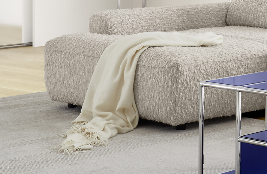 Teilansicht eines modernen Wohnzimmers mit einem strukturierten, hellen Sofa, über dessen Armlehne eine cremefarbene Decke gelegt ist, die teilweise auf dem Boden liegt. Das Sofa wirkt weich und gemütlich. Im Vordergrund steht ein blauer, glänzender Couchtisch mit Chromgestell. Der Boden ist mit einem hellen Teppich bedeckt, im Hintergrund sind ein Holzfußboden und eine weiße Wand zu erkennen. Das Bild betont ein zeitgemäßes, behagliches Design mit Fokus auf Textur und Farbkontraste.