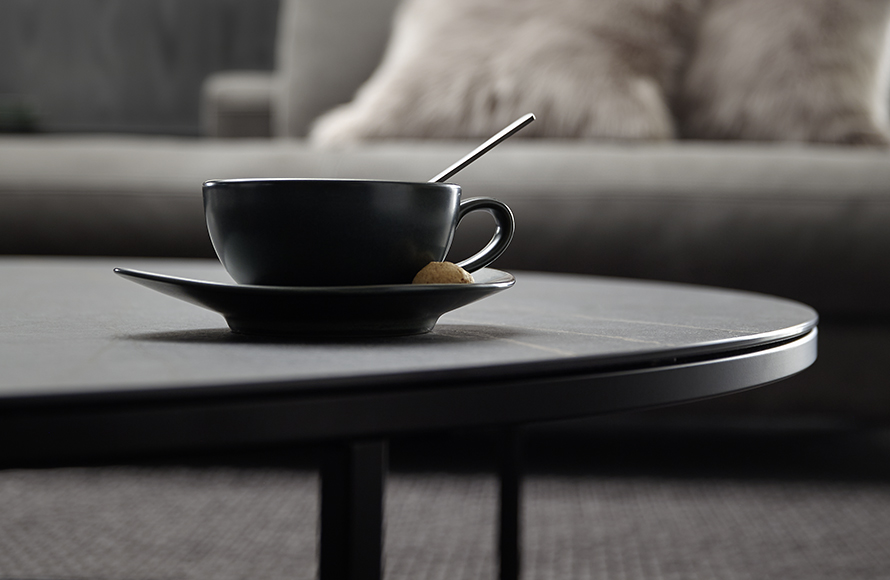 Nahaufnahme einer schwarzen Tasse mit Untertasse und Löffel auf einem runden Tisch. Auf der Untertasse liegt ein kleines Gebäck oder Keks neben der Tasse. Im Hintergrund ist unscharf ein Sofa mit einem flauschigen Kissen zu erkennen, was eine gemütliche und einladende Atmosphäre vermittelt.