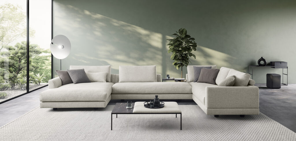 Modernes Wohnzimmer mit einem großen, beigefarbenen Ecksofa, dekoriert mit grauen und beigen Kissen. Vor dem Sofa steht ein niedriger rechteckiger Couchtisch mit dunkler Platte und schwarzen Beinen, auf dem ein Tablett mit Dekorationen liegt. Links befindet sich eine große Stehlampe mit rundem weißen Schirm. Ein bodentiefes Fenster bietet Blick auf einen grünen Garten. Rechts steht ein Konsolentisch mit Dekorationsobjekten wie einer schwarzen Vase und einer kleinen Box, daneben eine hohe grüne Pflanze. Der Raum ist mit einem hellgrauen Teppich ausgelegt und die Wände sind in einem sanften Grünton gestrichen. Das Design ist minimalistisch und modern, mit neutralen Farben und viel natürlichem Licht.
