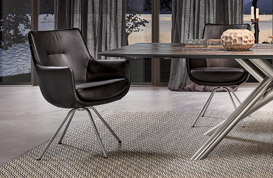 Modernes Interieur mit einem Essbereich, bestehend aus zwei schwarzen Lederstühlen mit Metallbeinen – einer im Vordergrund, der andere teilweise hinter einem Esstisch sichtbar. Der Tisch hat eine dunkle Oberfläche und gekreuzte Metallbeine. Darauf stehen ein strukturierter, kugelförmiger Dekogegenstand und ein kleines rechteckiges Glasgefäß. Im Hintergrund sind große Fenster mit Vorhängen zu sehen, die den Blick auf eine ruhige Außenlandschaft mit Wasser und Bäumen freigeben. Der Boden ist mit einem gemusterten Teppich bedeckt.