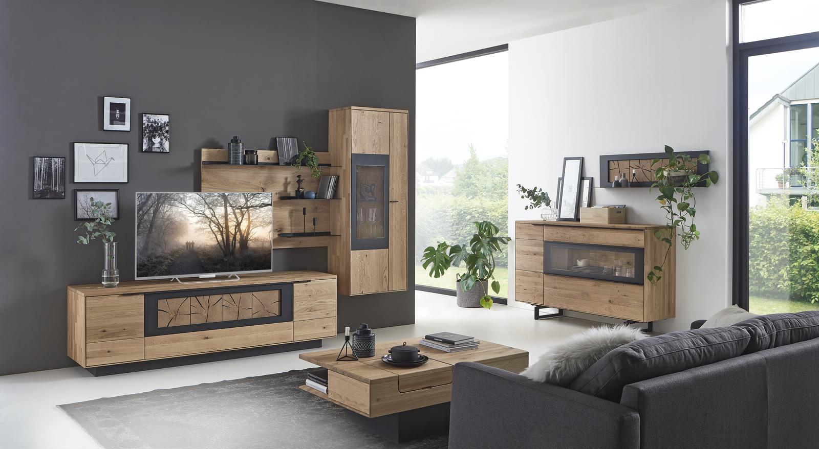 Modernes Wohnzimmer mit stilvollen Möbeln in Holz und Schwarz. Zu sehen sind ein großes TV-Board mit Flachbildfernseher, eine wandhängende Vitrine, ein Sideboard und ein Couchtisch mit Dekoration. Dunkle Akzentwand mit Bildern und Regal, helle Fensterfront und grüne Pflanzen sorgen für eine gemütliche, lichtdurchflutete Atmosphäre.