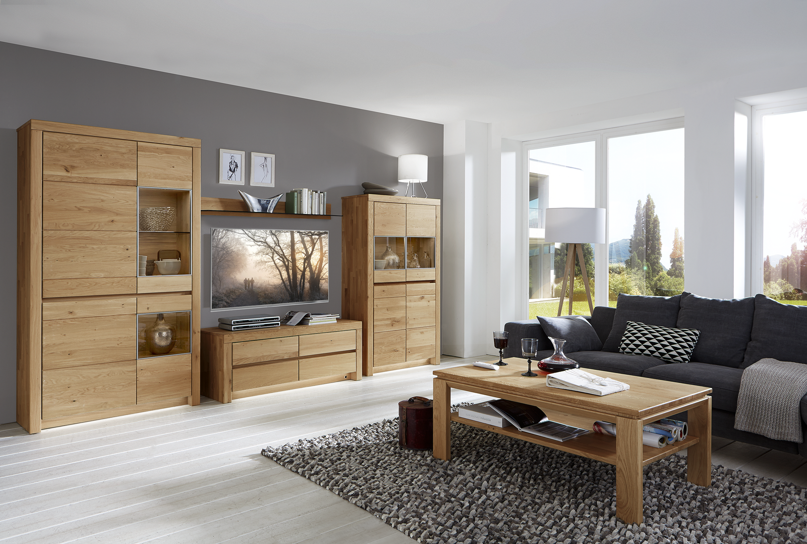 Modernes Wohnzimmer mit großem Holz-Entertainment-Center, Flachbildfernseher, grauem Ecksofa und Couchtisch. Dekorative Elemente wie Bücher, Decken und eine Stehlampe sorgen für wohnliches Flair. Große Fenster bringen Tageslicht und bieten Blick ins Grüne.