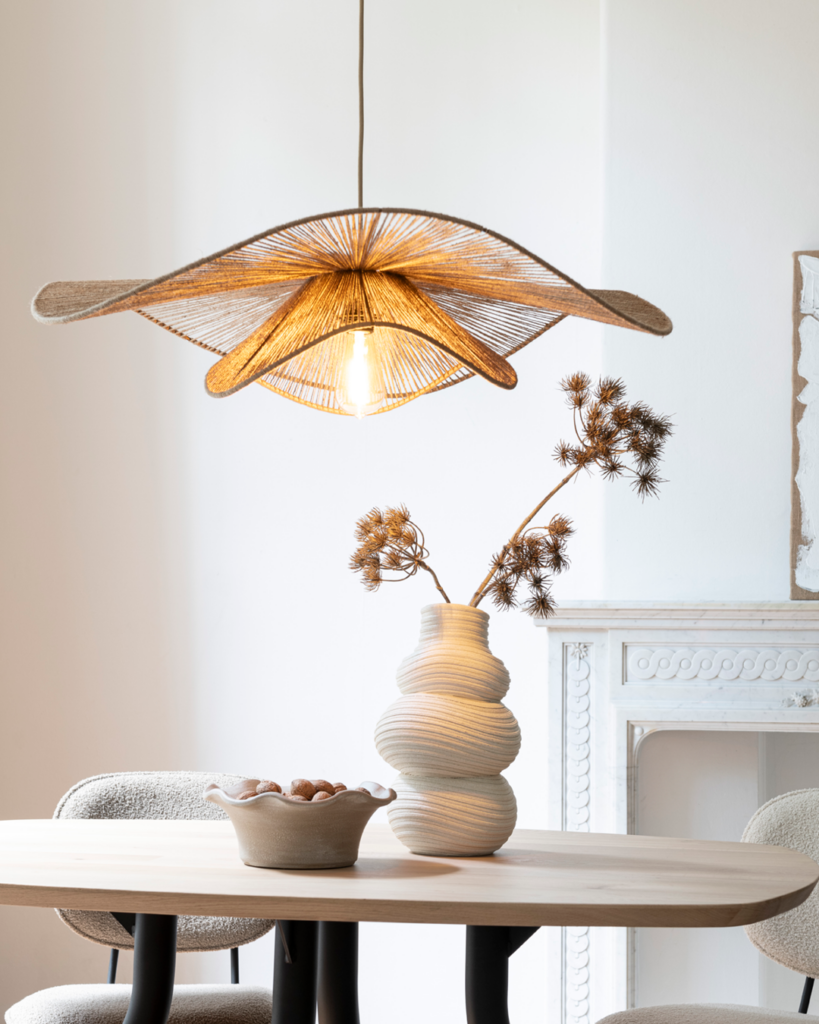 Moderne Wohnszene mit einem auffälligen, hängenden Leuchtkörper, einem Holztisch und dekorativen Elementen. Die Lampe besteht aus geflochtenem Material in sternförmigem Muster und strahlt warmes Licht aus. Darunter steht ein Holztisch mit schwarzen Beinen. Auf dem Tisch befinden sich eine strukturierte Keramikvase mit einem Arrangement aus Trockenpflanzen sowie eine kleine Schale mit Nüssen. Im Hintergrund ist ein weißer Kamin mit kunstvollen Verzierungen zu sehen. Die Kombination aus natürlichen Materialien und modernen Formen schafft eine gemütliche und stilvolle Atmosphäre.