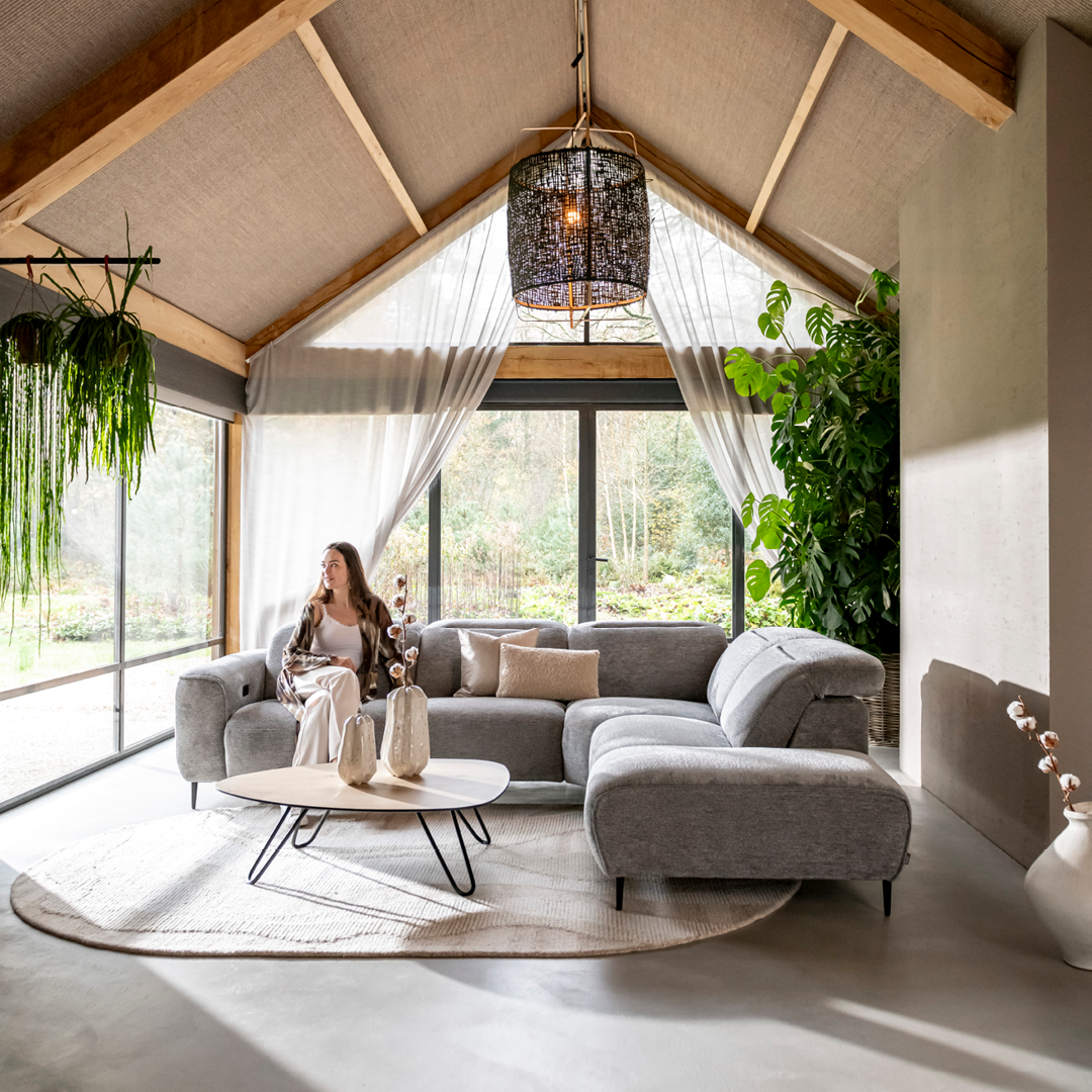 Modernes, gemütliches Wohnzimmer mit hoher Decke und sichtbaren Holzbalken, großen Fenstern mit Blick ins Grüne, grauem Ecksofa, weißem Couchtisch, dekorativen Vasen und Pflanzen, sowie einem schwarzen Hängelicht – stilvoll und lichtdurchflutet eingerichtet.
