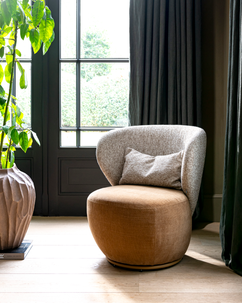 Modernes Wohnzimmer mit einem rund geformten Sessel in Braun und Grau, dekorativem Kissen, großem grünen Zimmerpflanze in einem Topf auf einem Buch, hellem Holzfußboden und einem Fenster mit schwarzen Rahmen und dunklen Vorhängen im Hintergrund.