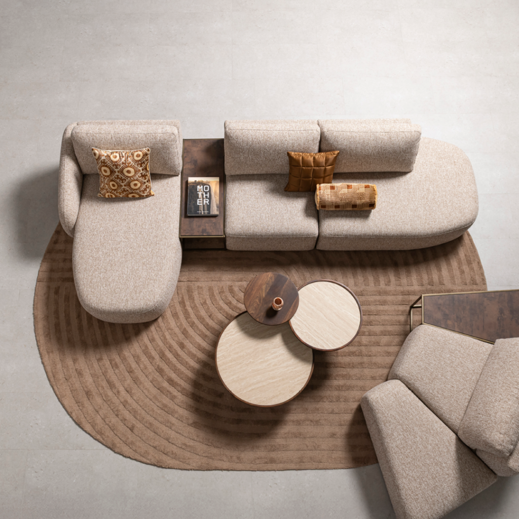 Vogelperspektive auf ein modern eingerichtetes Wohnzimmer mit beigem Ecksofa, rundem Couchtisch, braunem Teppich mit Kreismuster und einem Buch mit dem Titel „MOTHER“ auf einem Beistelltisch.