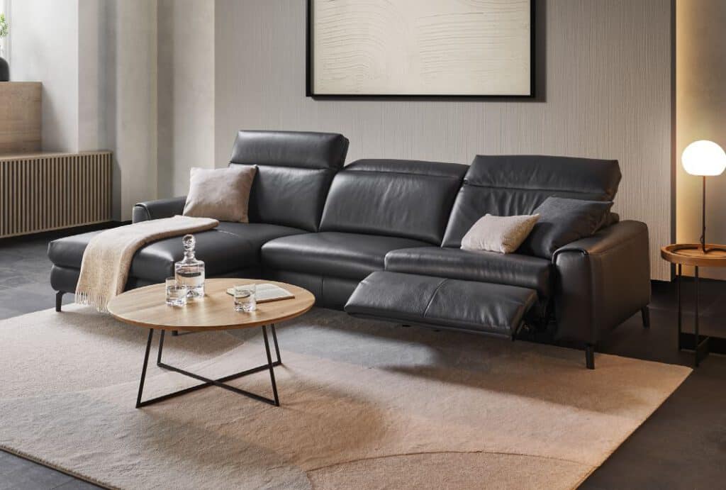 Modernes Wohnzimmer mit dem WK 660 VENOSA Relax-Sofa in dunkelgrauem Echtleder. Elektrisches Fußteil ausgefahren, dekoriert mit hellen Kissen und Decke. Runde Holz-Couchtische, stilvolle Wandkunst und warme Beleuchtung schaffen eine elegante Wohlfühlatmosphäre.