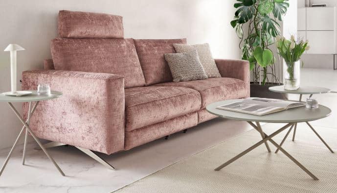 Wohnzimmer mit dem WK 605 VARIATIONE 2-Sitzer Sofa in roséfarbenem Stoff. Zwei Zierkissen, runde Beistelltische mit Blumen, Buch und Lampe. Helle Teppiche, grüne Pflanze und moderne Einrichtung betonen den gemütlich-stylischen Charakter.