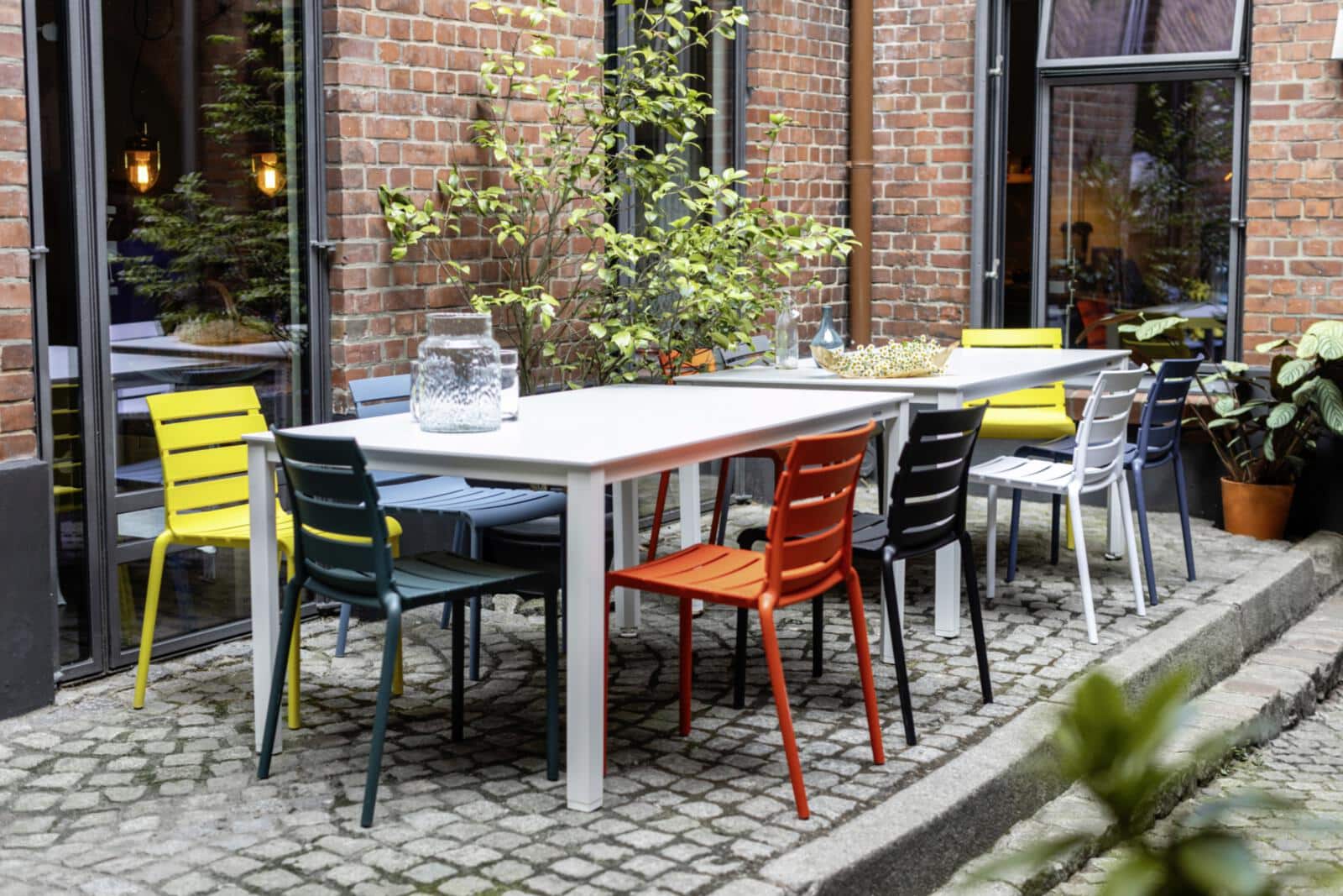 Zwei weiß gedeckte Tische stehen in einem charmanten Innenhof mit Kopfsteinpflaster, umrahmt von Backsteinwänden und üppigem Grün. Bunte Stühle in Gelb, Blau, Grün und Orange laden zum Verweilen ein – ein lebendiger Ort voller Farbe, Geselligkeit und urbaner Gemütlichkeit.