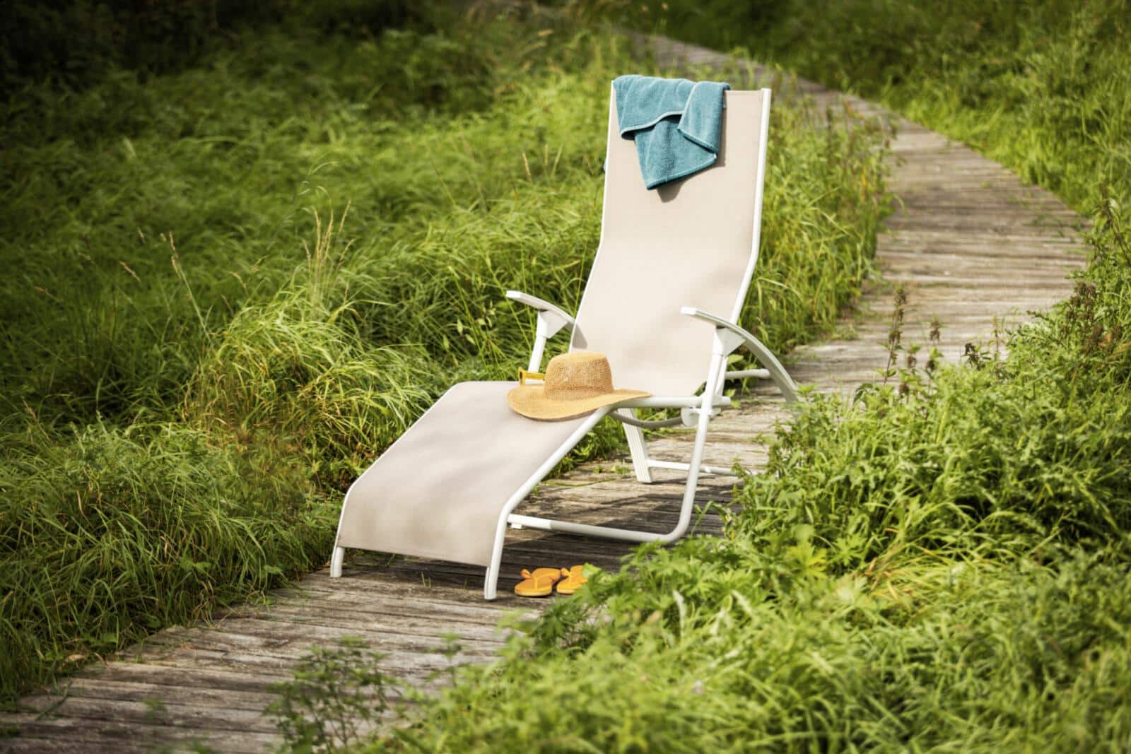 Eine beige Liege mit Strohhut, Sonnencreme und Handtuch steht auf einem Holzsteg im Grünen – daneben liegen orangefarbene Flip-Flops. Ein stiller Moment voller Sommer, Leichtigkeit und natürlicher Entspannung.