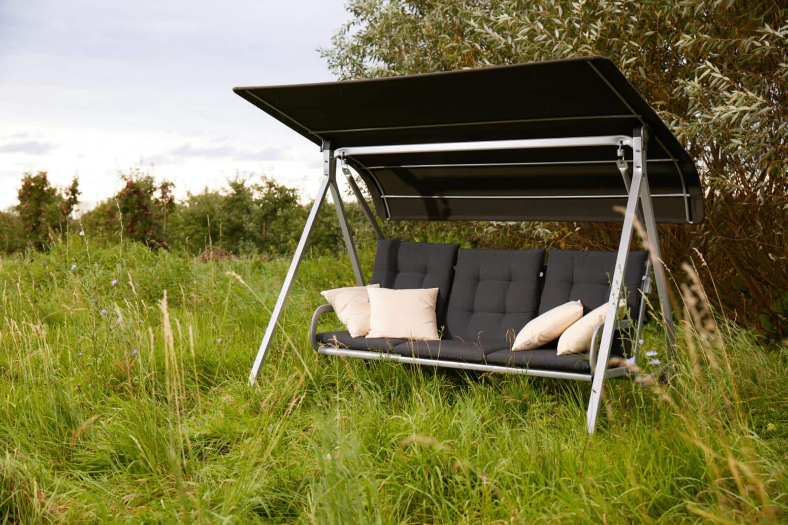 Eine moderne Gartenschaukel mit dunklem Sonnendach und weichen Polstern steht auf einer Wiese, umgeben von hohen Gräsern und Bäumen – ein ruhiger Rückzugsort, der Design und Natur harmonisch verbindet.