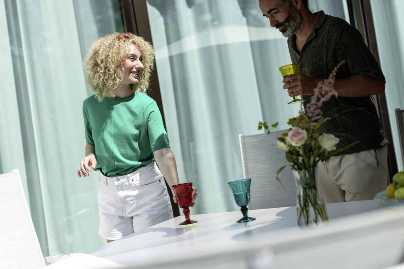 Zwei Personen stehen entspannt an einem weiß gedeckten Tisch mit bunten Gläsern, frischen Blumen und einer Schale voller grüner Äpfel und Zitronen – Sonnenlicht fällt durch transparente Vorhänge und schafft eine sommerlich-leichte Atmosphäre. Ein Moment voller Farbe, Frische und Geselligkeit.
