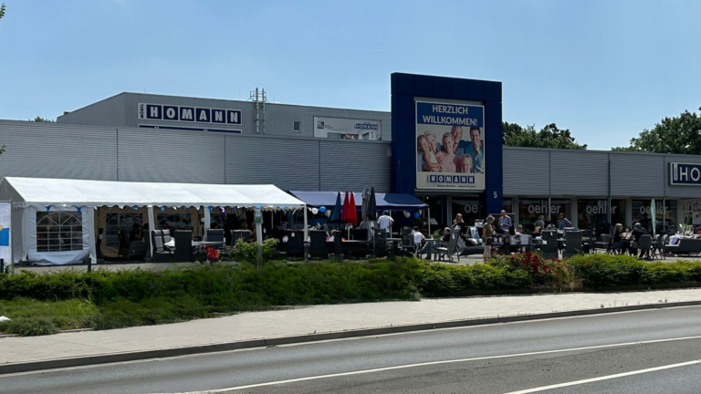 Außenansicht eines festlich dekorierten Möbelhauses mit dem Schriftzug „HOMANN“. Vor dem Gebäude stehen ein weißes Festzelt, Sitzgelegenheiten und mehrere Personen, die an einem Event teilnehmen. Ein großes Banner mit der Aufschrift „HERZLICH WILLKOMMEN!“ begrüßt die Gäste. Die Szene spielt sich bei sonnigem Wetter ab, mit klarer Sicht auf Straße, Fußweg und Begrünung.