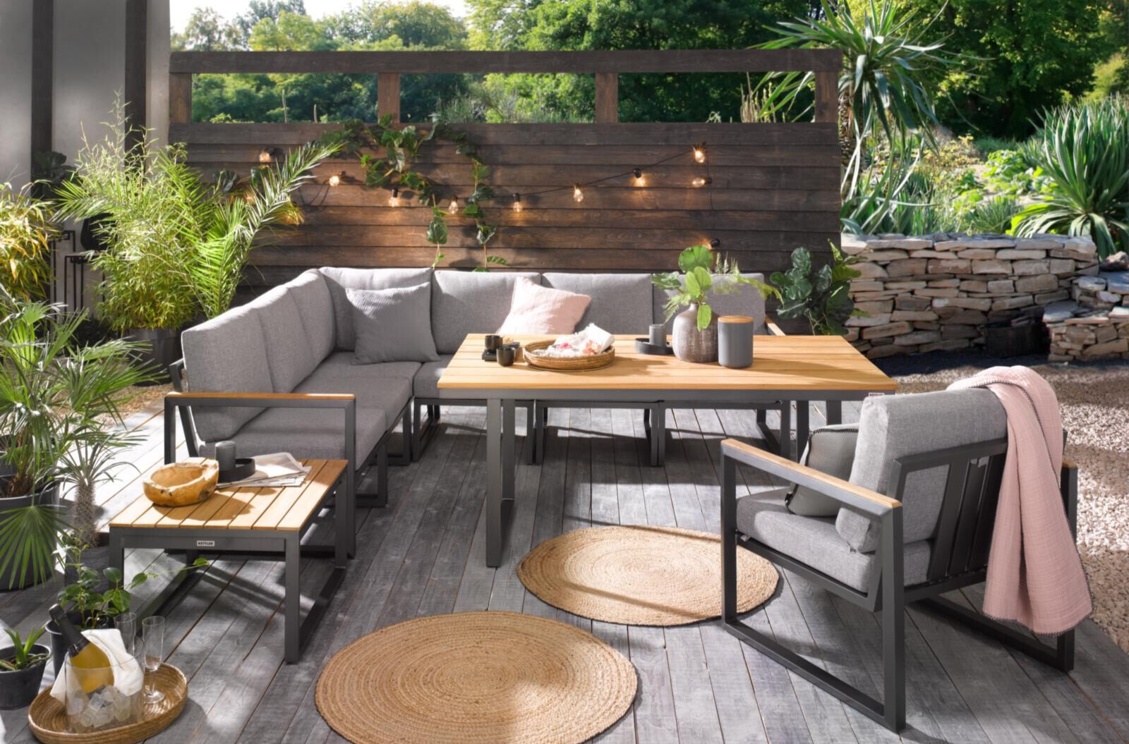 Stilvoll eingerichtete Terrasse mit grauen Polstermöbeln, Holztisch und liebevollen Details – umgeben von Pflanzen, Lichterketten und Naturstein entsteht ein moderner Outdoor-Wohnraum voller Gemütlichkeit und Designharmonie.