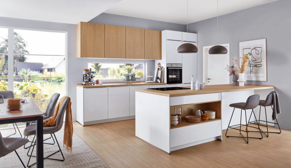 Moderne Küche mit weißer und heller Holzoptik, zentraler Kochinsel und gemütlichem Essbereich. Zwei Barhocker stehen an der Insel, darüber hängen dunkle Pendelleuchten. Durch das große Fenster fällt Tageslicht auf die stilvolle Einrichtung mit offenen Regalen, Deko und Blick ins Grüne. Die Szene zeigt ein einladendes, harmonisches Wohngefühl – perfekt für Inspiration beim Küchenkauf.
